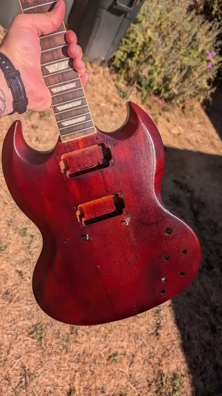 A person holds a red electric guitar body outdoors, showing its unfinished state with exposed wood, missing pickups, and…