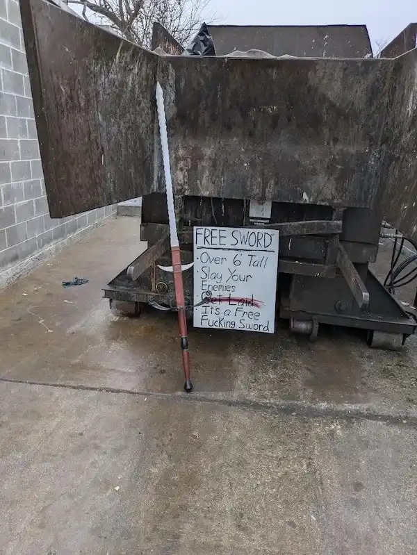 A large, rusty metal dumpster sits outdoors on a concrete surface, with a long sword leaning against its side. A handwritten…