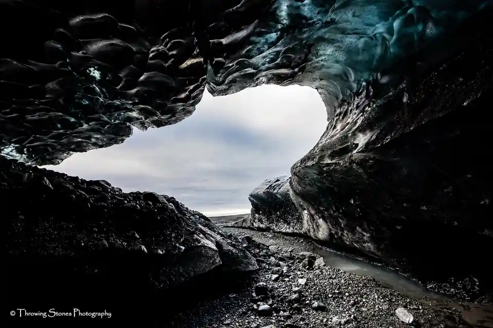A stunning photograph of a natural ice cave with a heart-shaped opening framing a cloudy sky and rocky terrain. The overlaid…