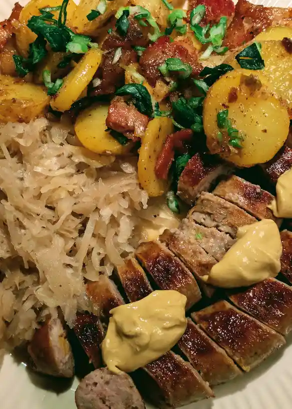 A close-up photo of a plate of food featuring sliced roasted meat with mustard dollops, sauerkraut, roasted potato slices…