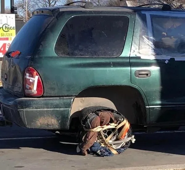 A person is curled up and seemingly asleep or resting on the ground next to the rear wheel of a green SUV, which has its…