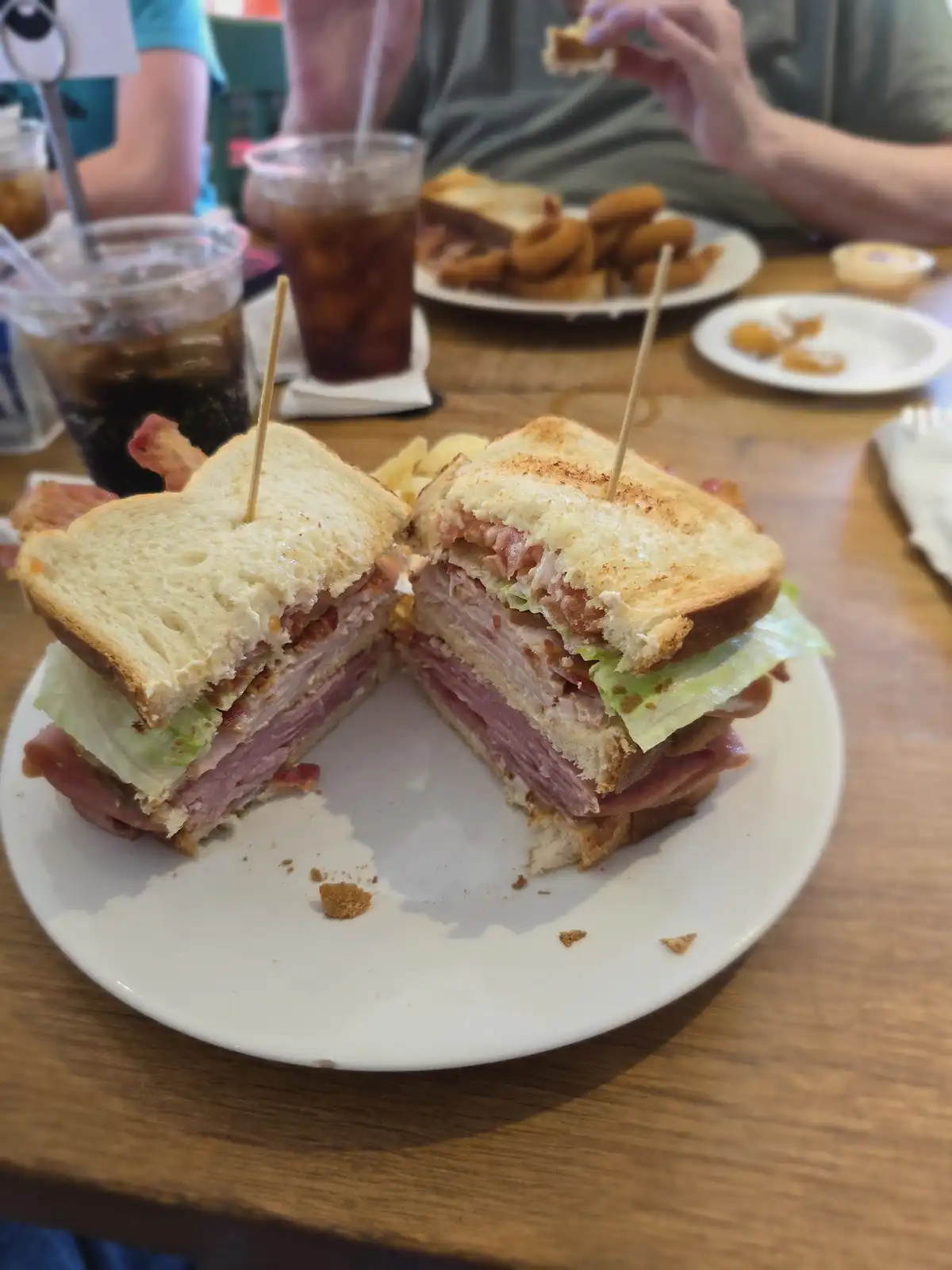 A large, layered club sandwich cut in half and held together with toothpicks sits on a white plate on a wooden table. In the…