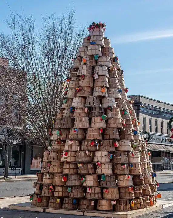 A large, cone-shaped structure made of stacked wooden baskets stands outdoors on a sidewalk, decorated with strings of…