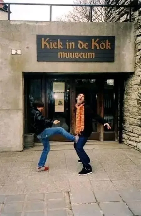 Two men are playfully kicking each other in front of a building with a sign that reads 'Kiek in de Kook muuseum'. One man is…