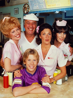 A group photo of the cast of 'The Facts of Life' from the 1980s, smiling and posing together in a diner setting. The…