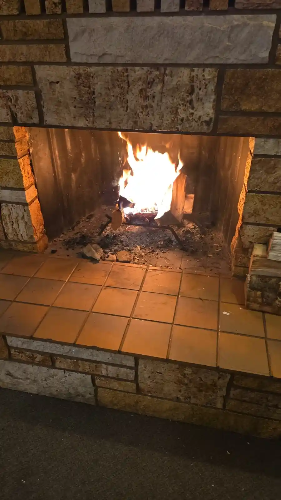 A close-up photo of a brick fireplace with a bright, active fire burning inside, casting warm light on the tiled hearth and…