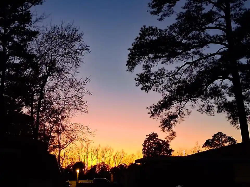 A serene sunset photo showing silhouetted trees and rooftops against a gradient sky transitioning from orange to purple. The…