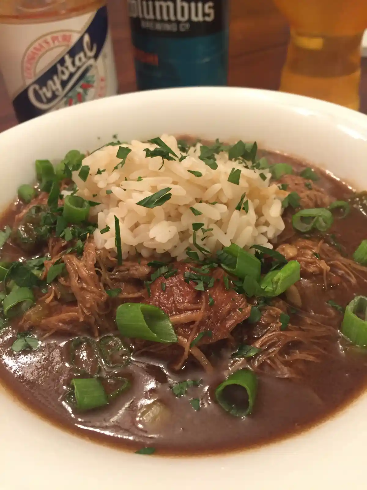 A close-up photo of a white bowl filled with a dark brown stew containing shredded meat, rice, chopped green onions, and…