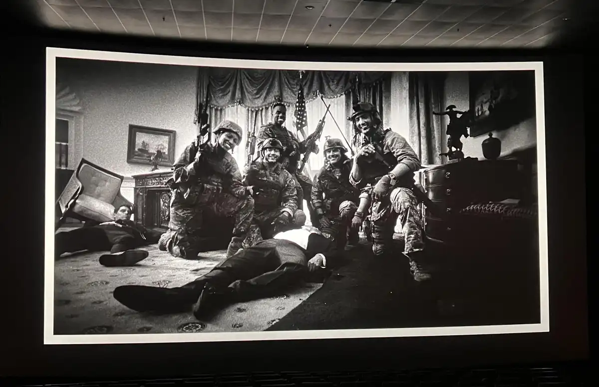 A black-and-white photo projected on a cinema screen shows five smiling soldiers in combat gear posing in a formal room with…