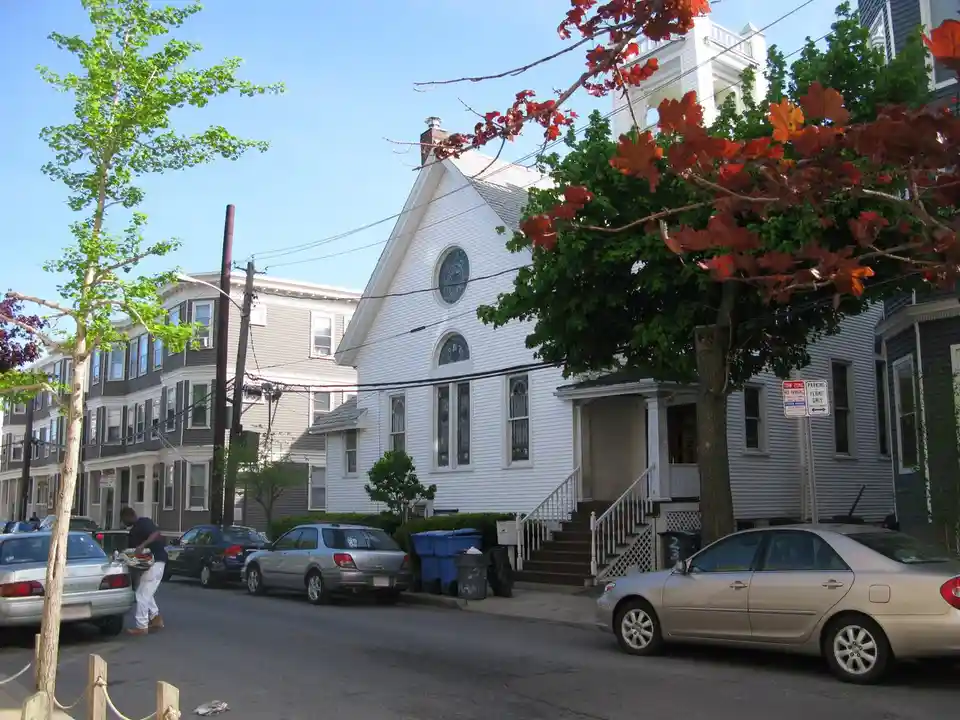 A street view shows a white church with stained glass windows and a bell tower, surrounded by parked cars and trees with red…