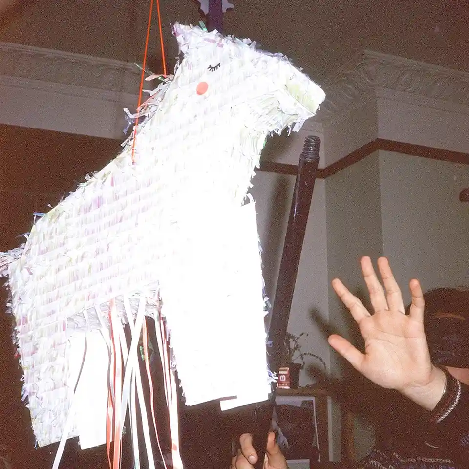 A person's hand is raised in front of a hanging, white, horse-shaped piñata with a smiling face, as if about to strike it…