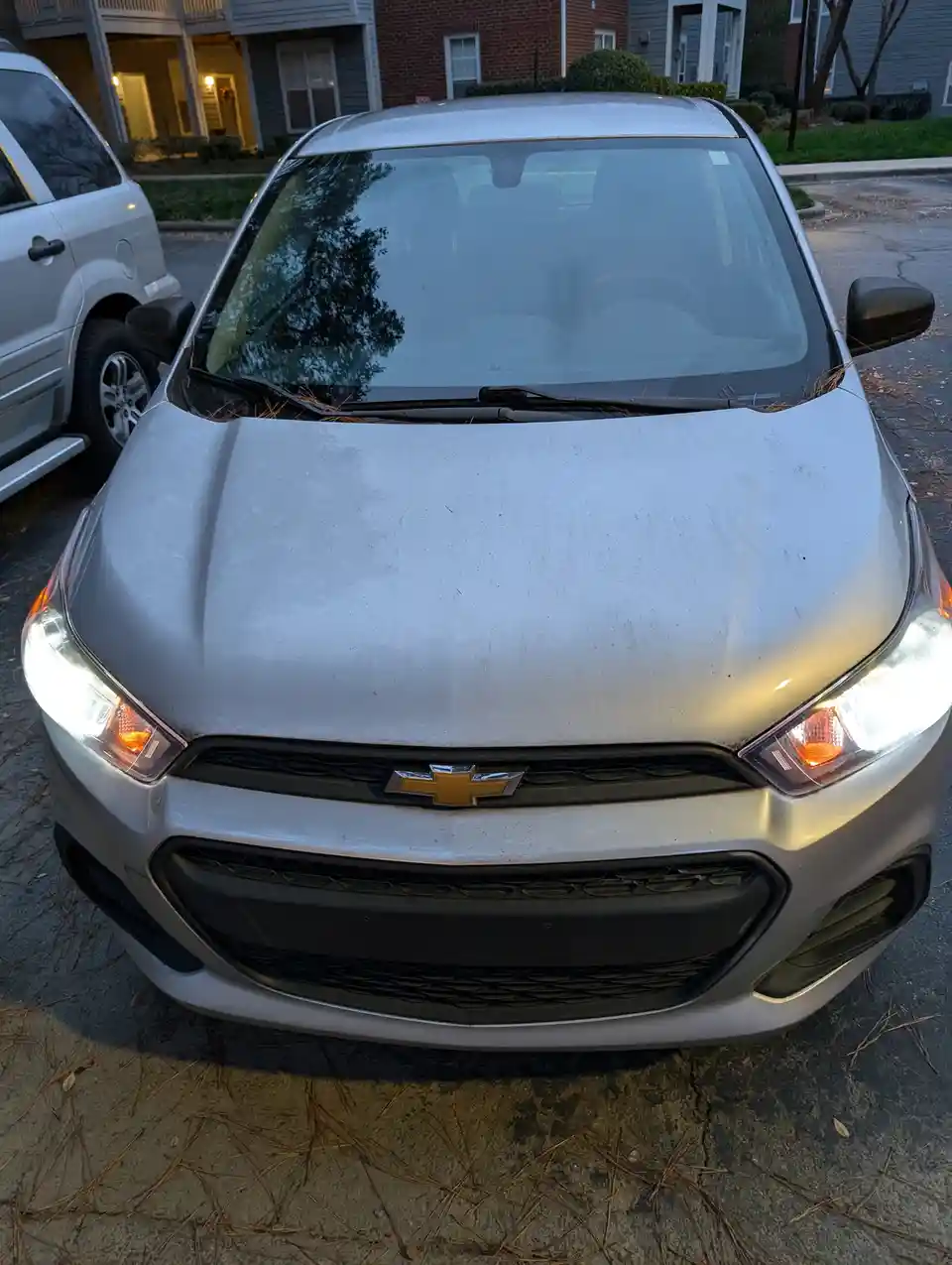 A front-facing photo of a silver Chevrolet car parked in a residential driveway, its hood and windshield covered in pine…