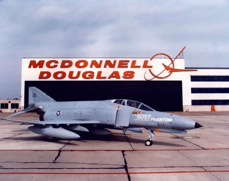 A gray military jet, marked '5057 LAST STAGGERS BUILT PHANTOM', is parked on tarmac in front of a large building with…