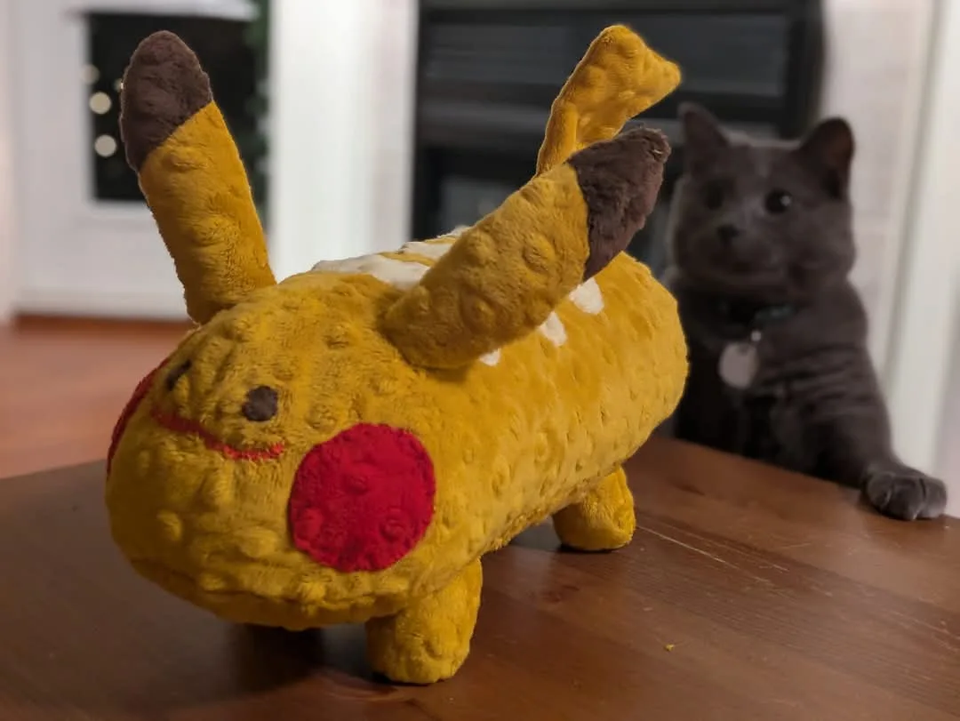 A plush Pikachu toy with a textured yellow body, red cheeks, and brown-tipped ears sits on a wooden table. In the blurred…