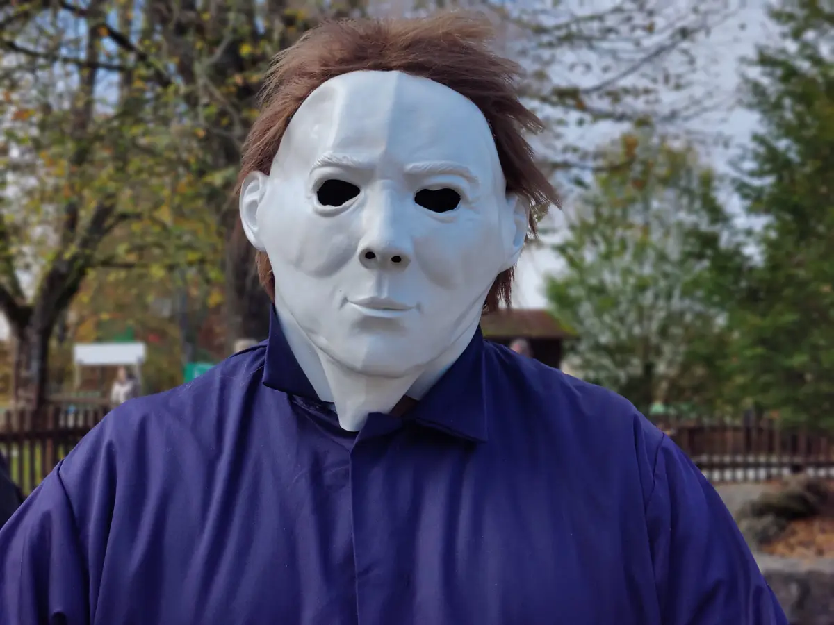 A person wearing a white Michael Myers mask and brown wig, dressed in a blue collared shirt, stands outdoors with trees and…
