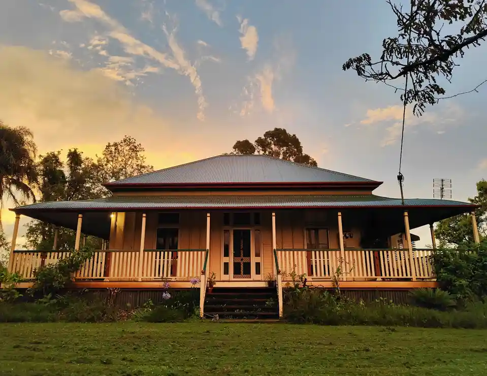 A serene, single-story house with a wide veranda and corrugated metal roof is pictured at sunset, surrounded by trees and…