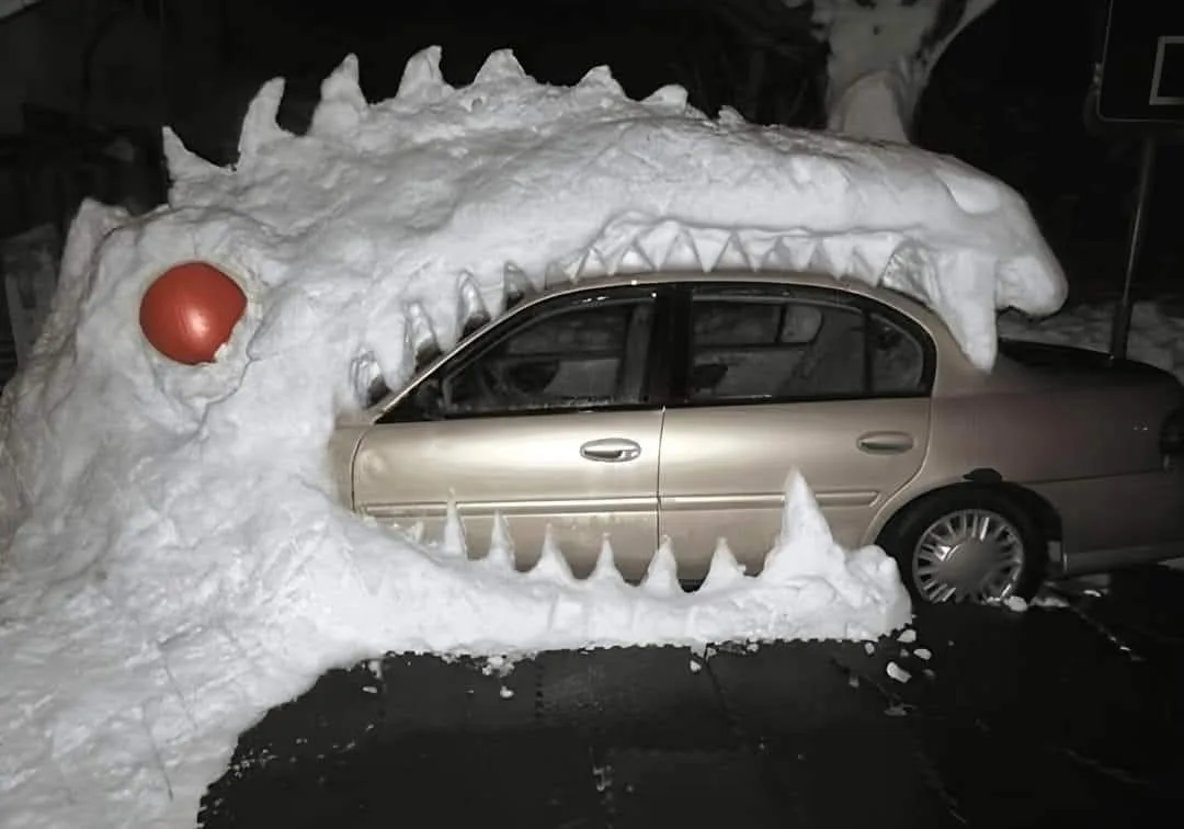 A gold sedan is partially buried under a large, detailed snow sculpture shaped like a dragon's head with sharp teeth and a…