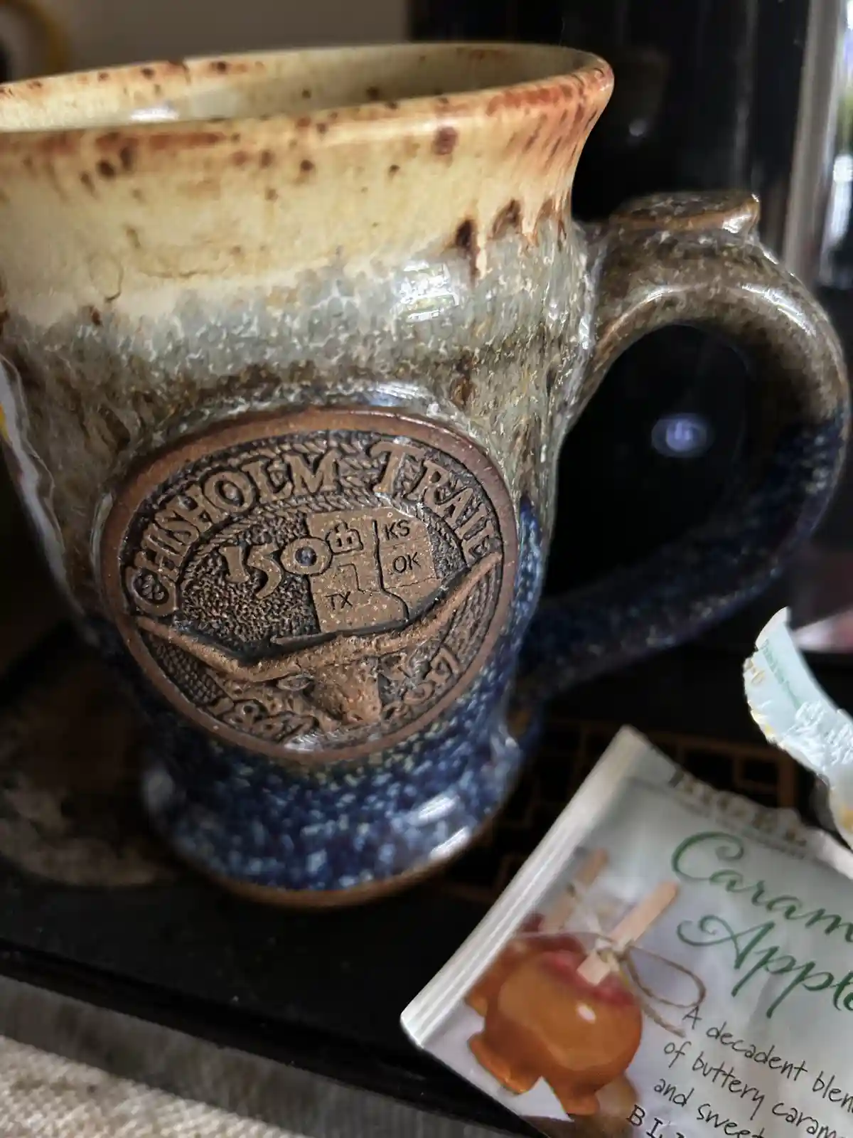 A rustic, speckled ceramic mug with a raised circular emblem reading 'CANYON TRAIL 150 TX KS OK' sits beside a packet of…
