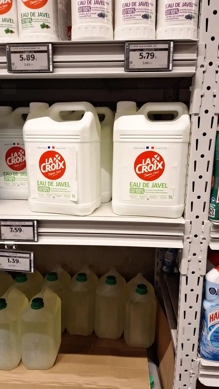 A supermarket shelf displays large white 5L jugs of 'La Croix Eau de Javel' (bleach) with price tags, humorously juxtaposed…