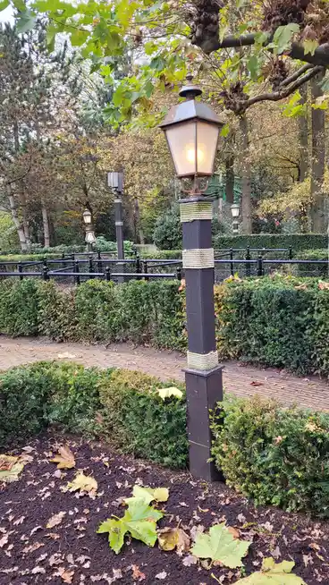 A photograph of a lit, vintage-style black lamppost wrapped with rope, standing in a park with autumn leaves on the ground…