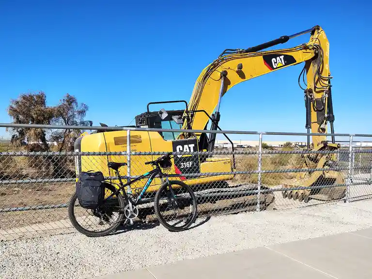 A black bicycle with a black pannier bag is leaning against a chain-link fence in front of a large yellow Caterpillar 316F L…