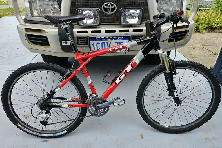 A red and black GT Zaskar Race mountain bike is leaning against the front bumper of a silver Toyota pickup truck with a…