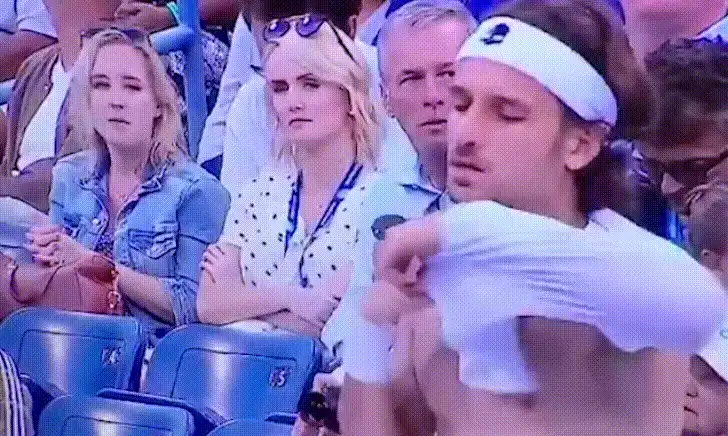 A shirtless man with a white headband removes his white shirt, revealing his bare torso, then holds up the shirt to his face…