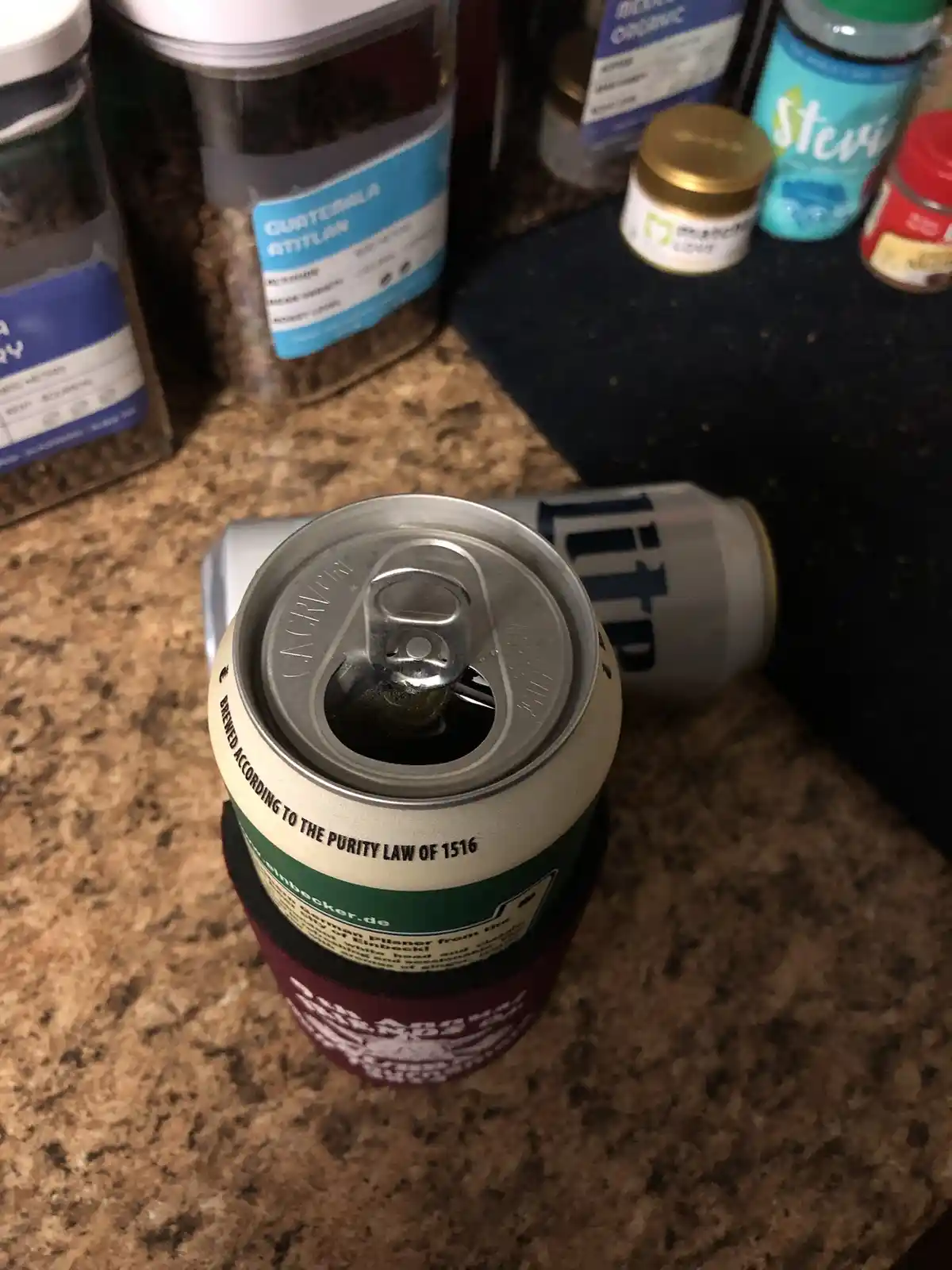 A close-up photo shows an open, upright beer can with a pull-tab, sitting on a countertop. Behind it, another can lies on…