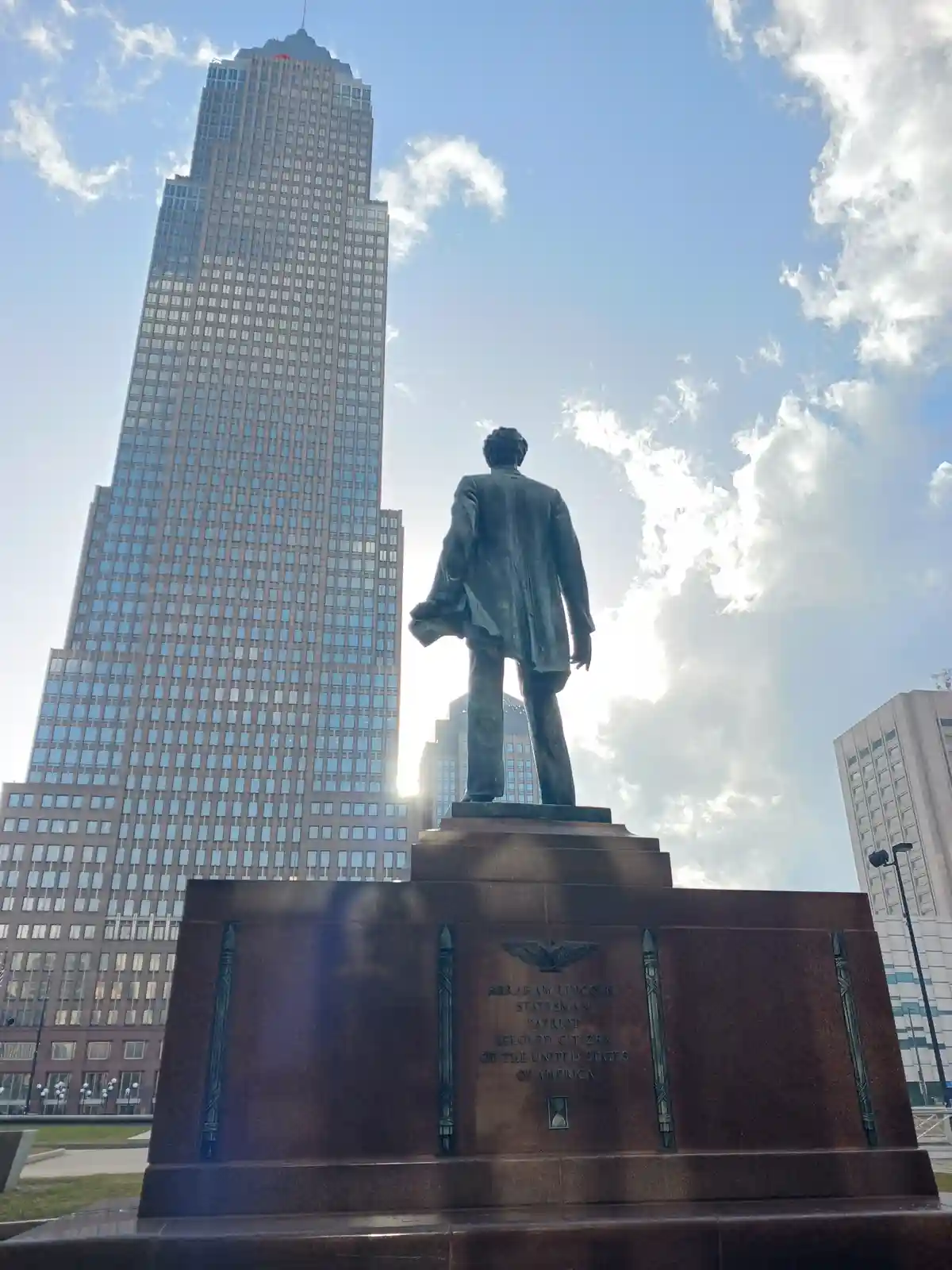 A low-angle photo shows the back of a bronze Abraham Lincoln statue standing on a granite pedestal with engraved text, set…