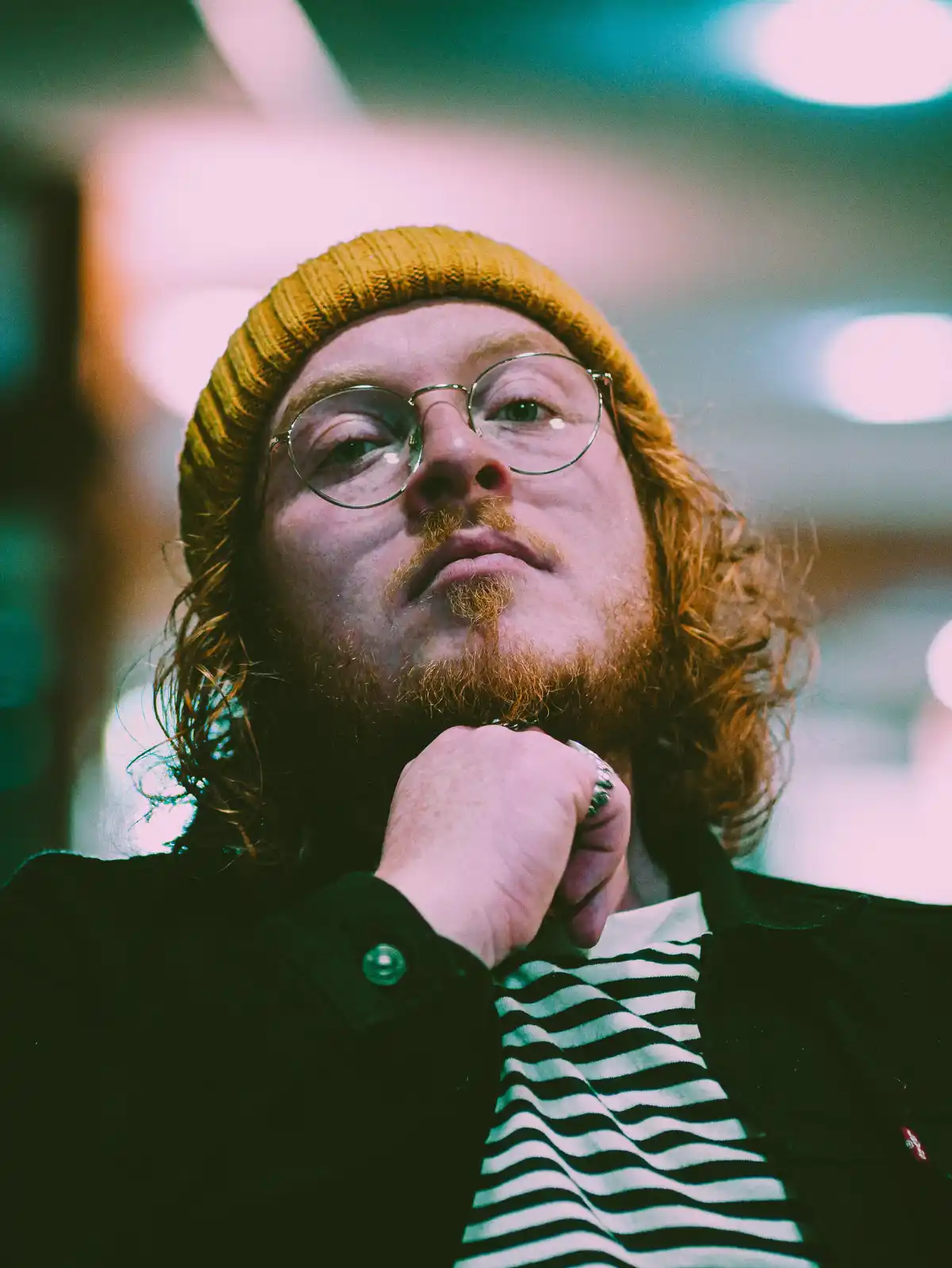 A man with red hair, a yellow beanie, and round glasses looks upward thoughtfully with his hand on his chin. He wears a…