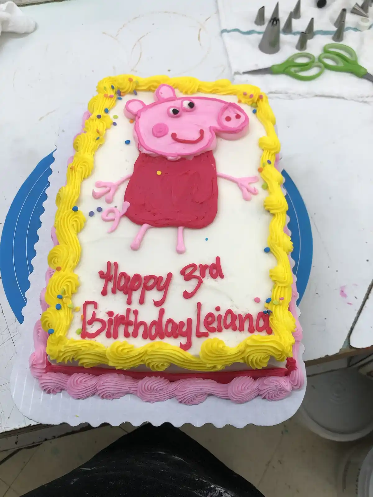 A rectangular birthday cake decorated with white frosting, a yellow piped border, and a fondant image of Peppa Pig in a red…