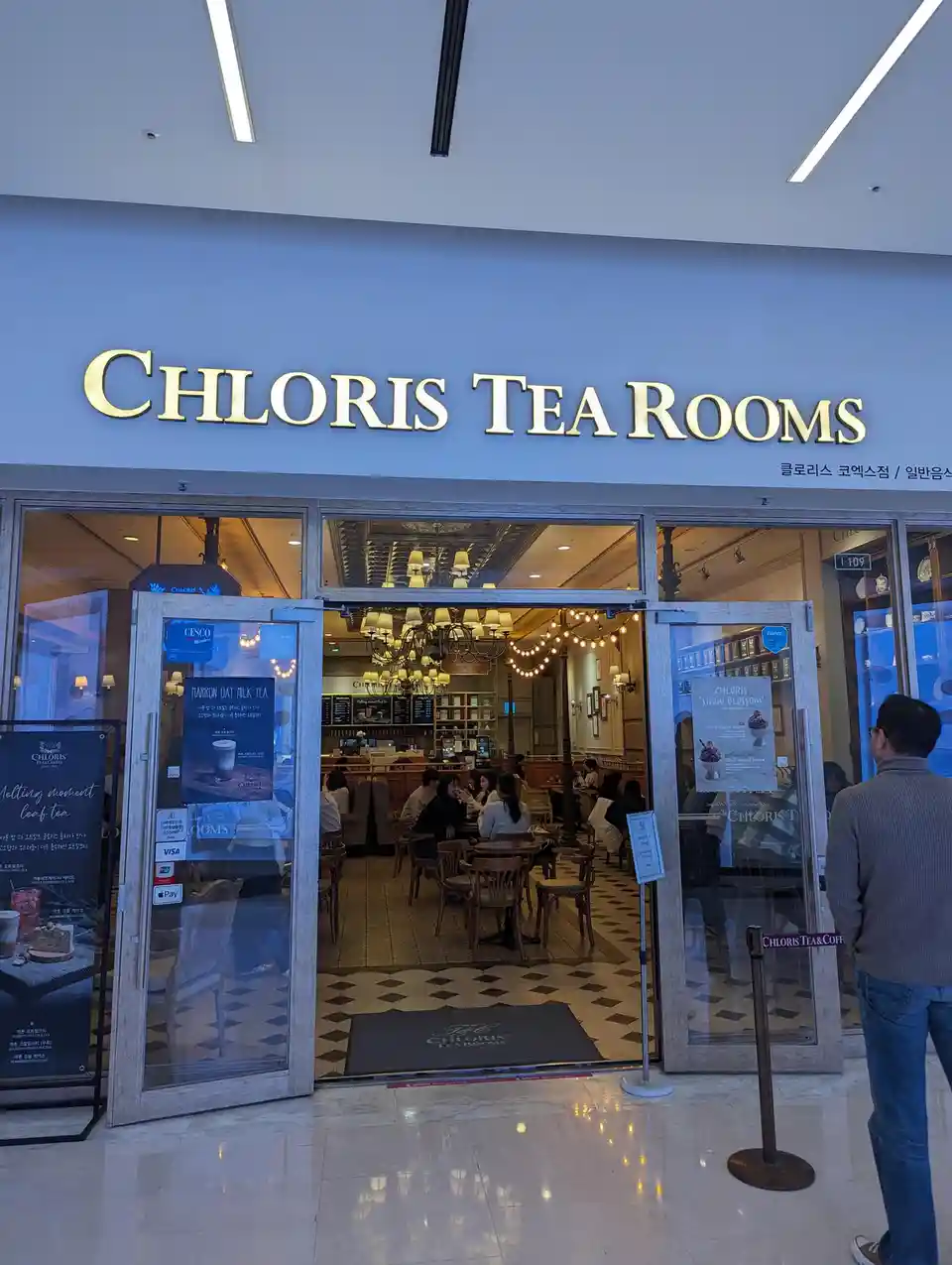 An exterior shot of a 'CHLORIS TEA ROOMS' storefront with glass doors open, revealing patrons seated inside under…