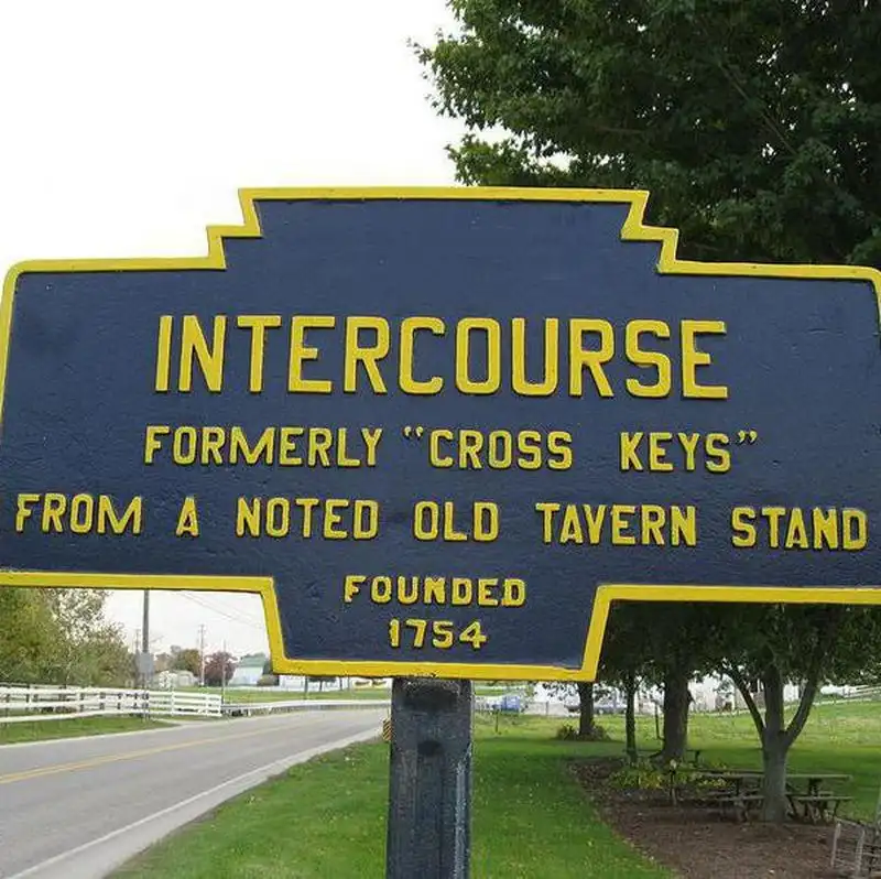 A blue historical marker sign with yellow text stands beside a road, identifying the town of 'INTERCOURSE' which was…