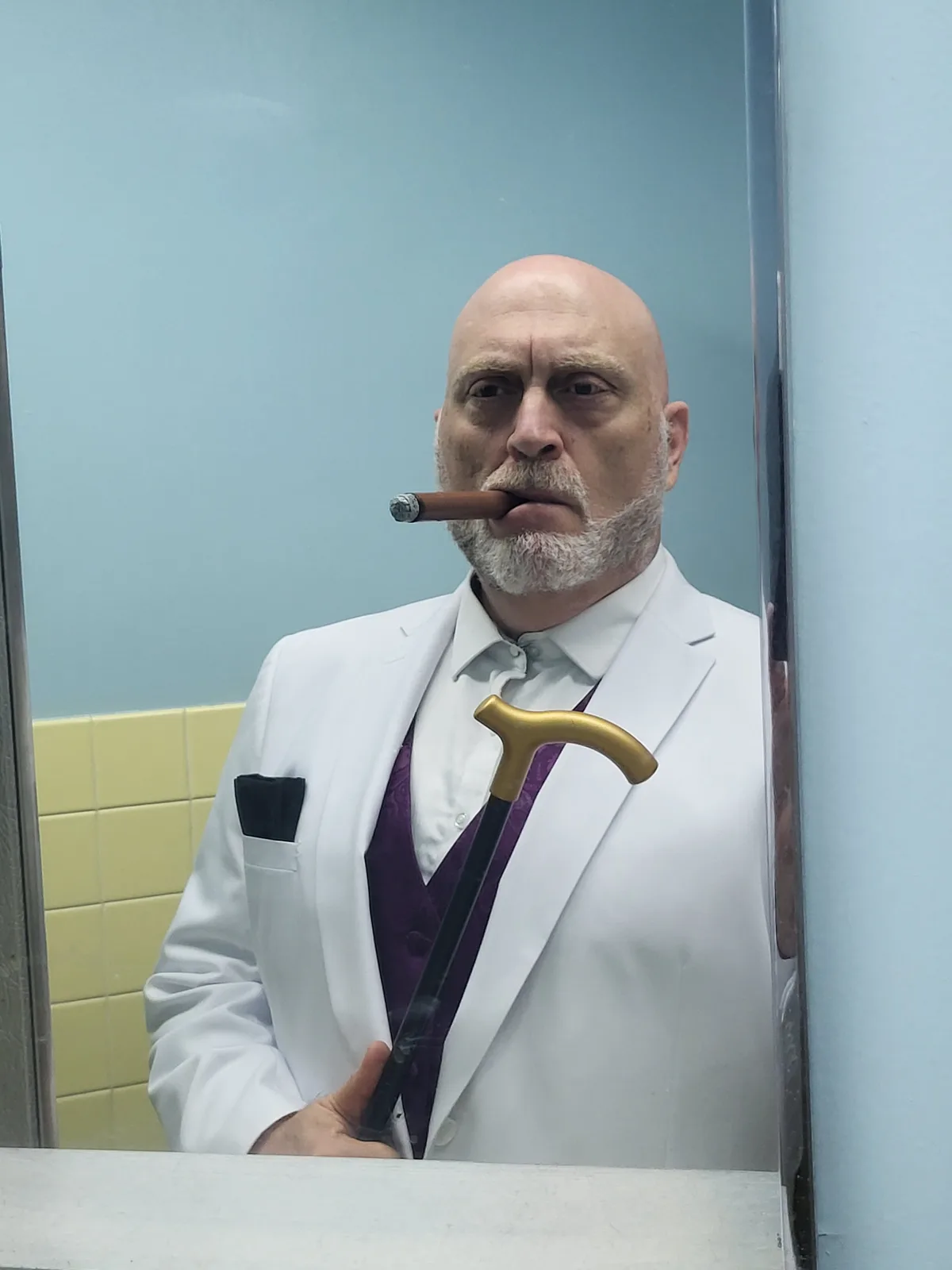 A bald man with a white beard stands in a bathroom mirror wearing a white lab coat over a purple vest and holding a…
