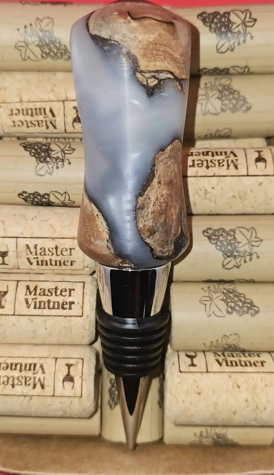 A wine stopper with a blue and brown wood-grain design stands upright in a stack of wine corks branded 'Master Vintner'. The…