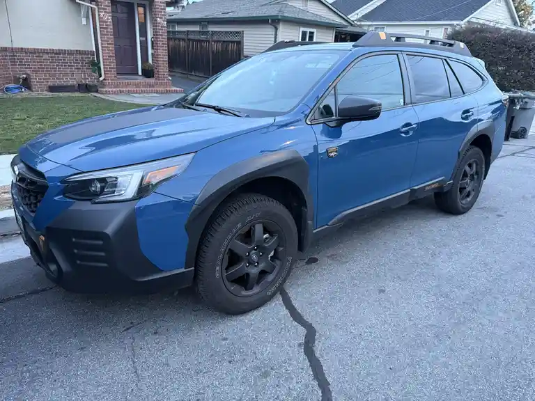 A blue Subaru Outback SUV with black trim and roof rails is parked on a residential street in front of a house. The car has…