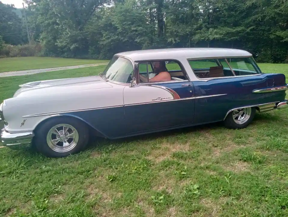 A man with a beard sits shirtless in the driver's seat of a vintage two-tone blue and silver station wagon parked on grass…