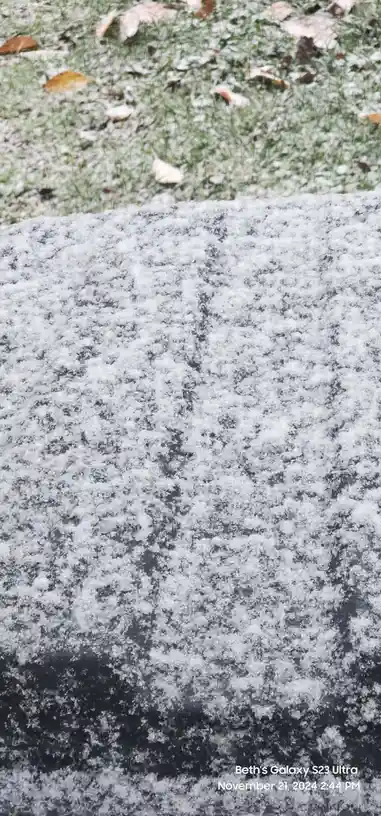 A close-up photo shows a dark, textured stone surface covered in a layer of frost, with frosty grass and fallen leaves…