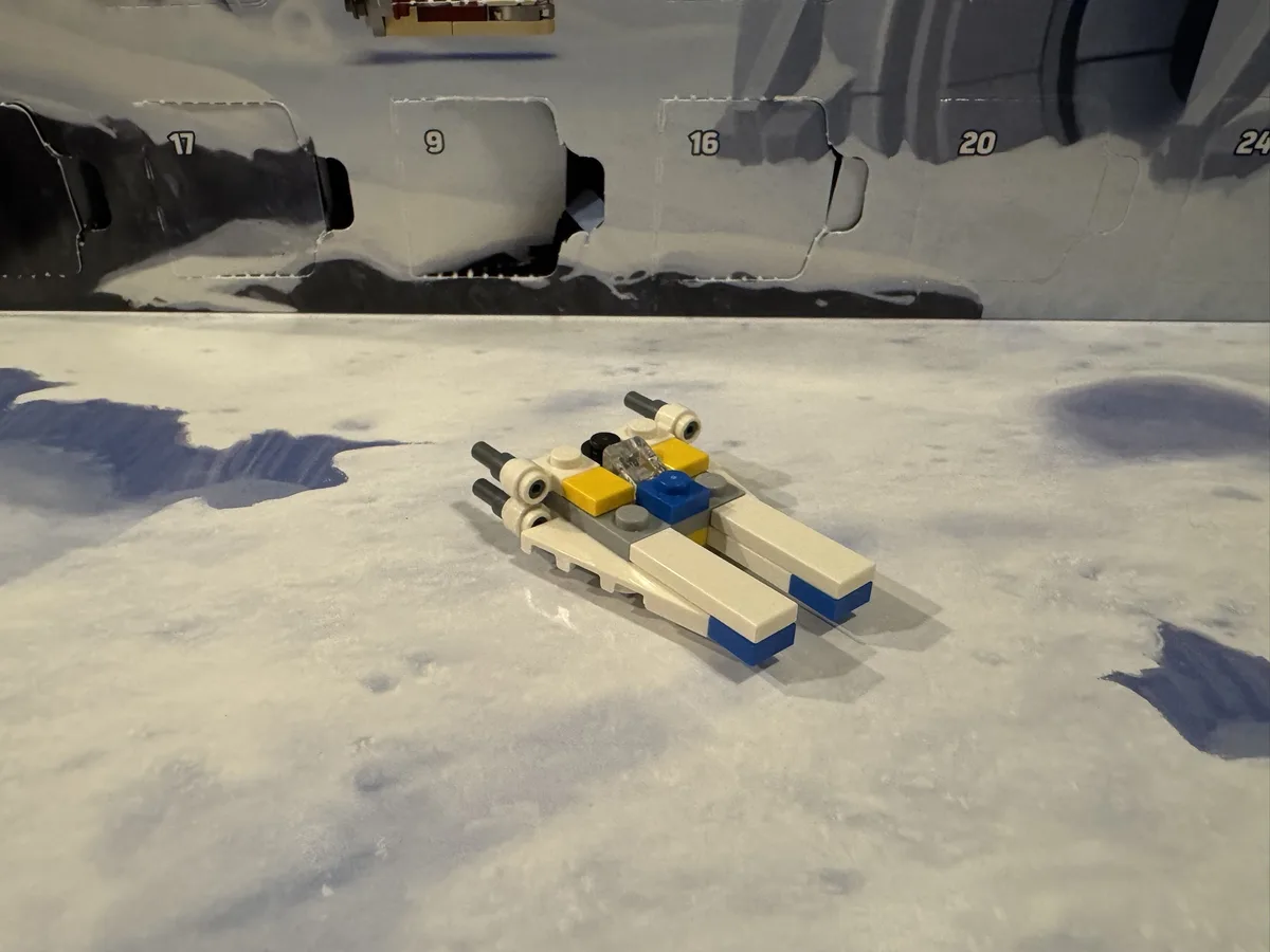A small, partially assembled LEGO X-wing fighter model sits on a snowy surface, in front of a torn LEGO advent calendar box…