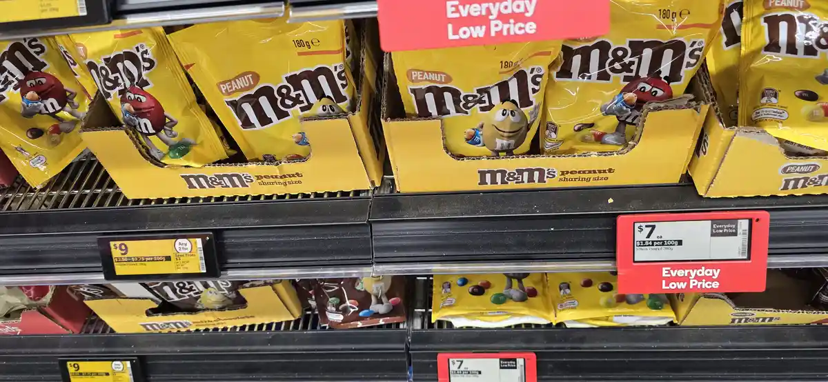 A supermarket shelf displays yellow bags of M&M's Peanut candy, each labeled 'sharing size' and priced at $7 each under an…