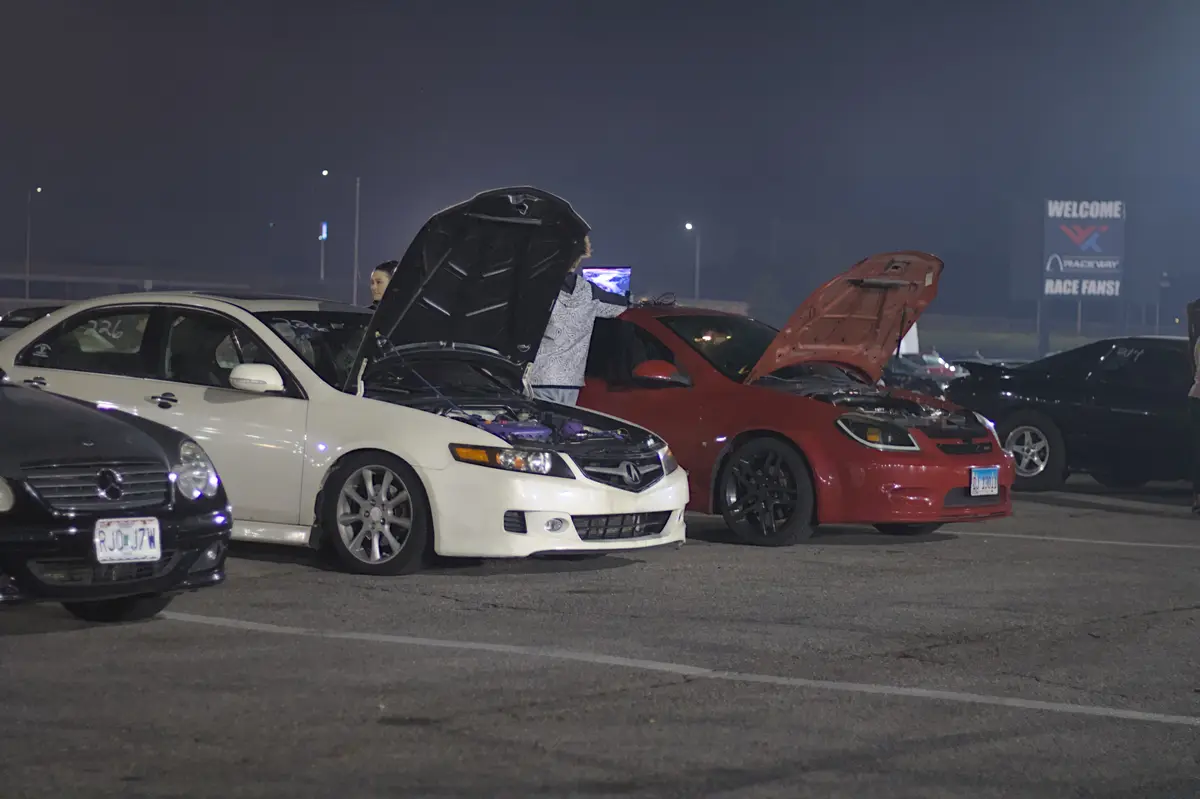 At night in a parking lot, a person in a patterned shirt stands between two cars with open hoods, holding a laptop. A woman…