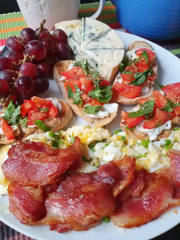 A plate of food featuring crispy bacon, scrambled eggs with green onions, tomato and basil bruschetta, a wedge of blue…