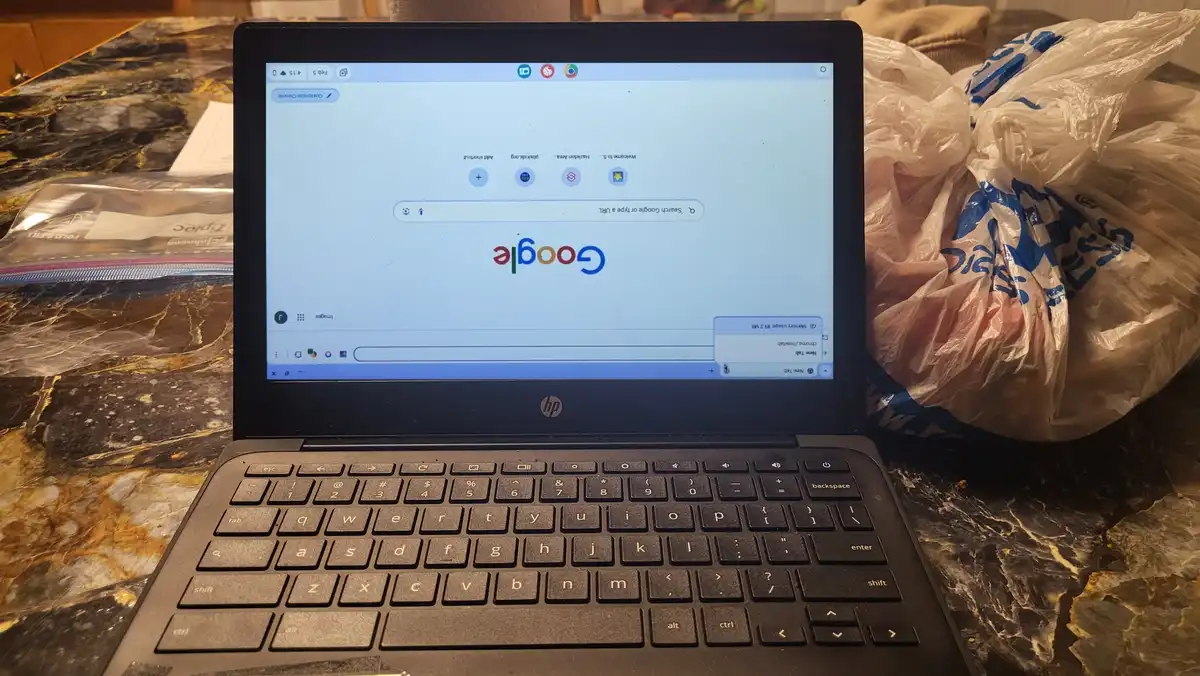 A laptop displaying the Google homepage sits on a marble countertop next to a crumpled plastic grocery bag. The screen shows…