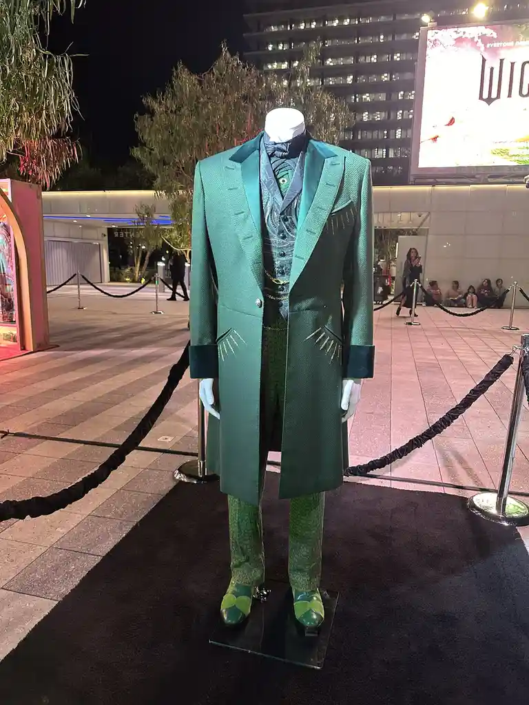 A full-body mannequin dressed in an elaborate, ornate green suit with matching pants and shoes, displayed outdoors at night…