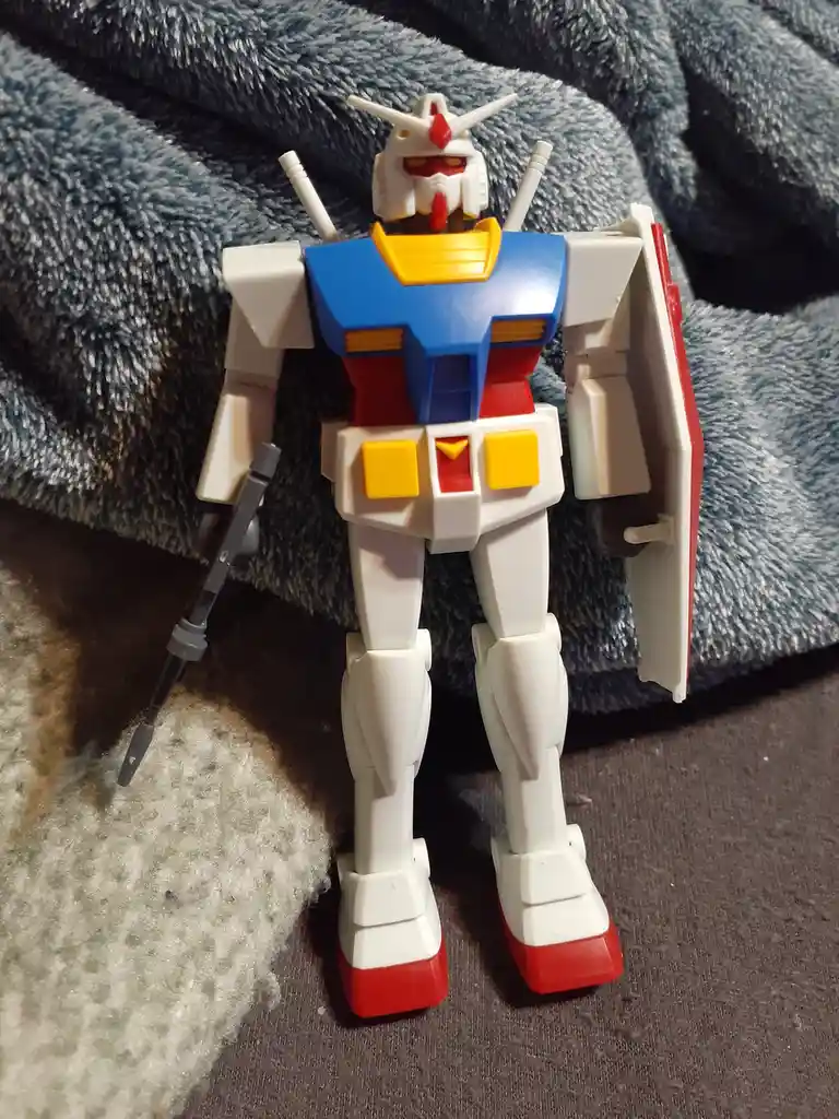 A white, blue, red, and yellow gundam robot toy stands upright on a dark carpet next to a blue textured blanket, holding a…