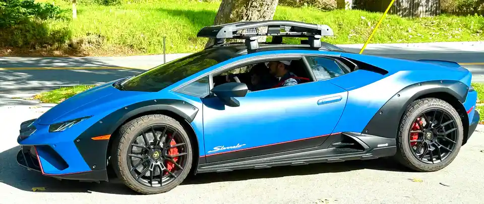 a blue lamborghini with black accents and red brake calipers is parked on a street, featuring a roof rack and all-terrain…