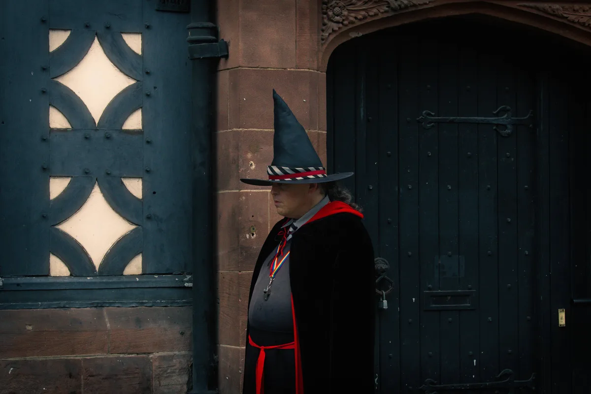 A man in a pointed witch hat and black robe stands solemnly beside ornate dark wooden doors. He wears a rainbow lanyard and…
