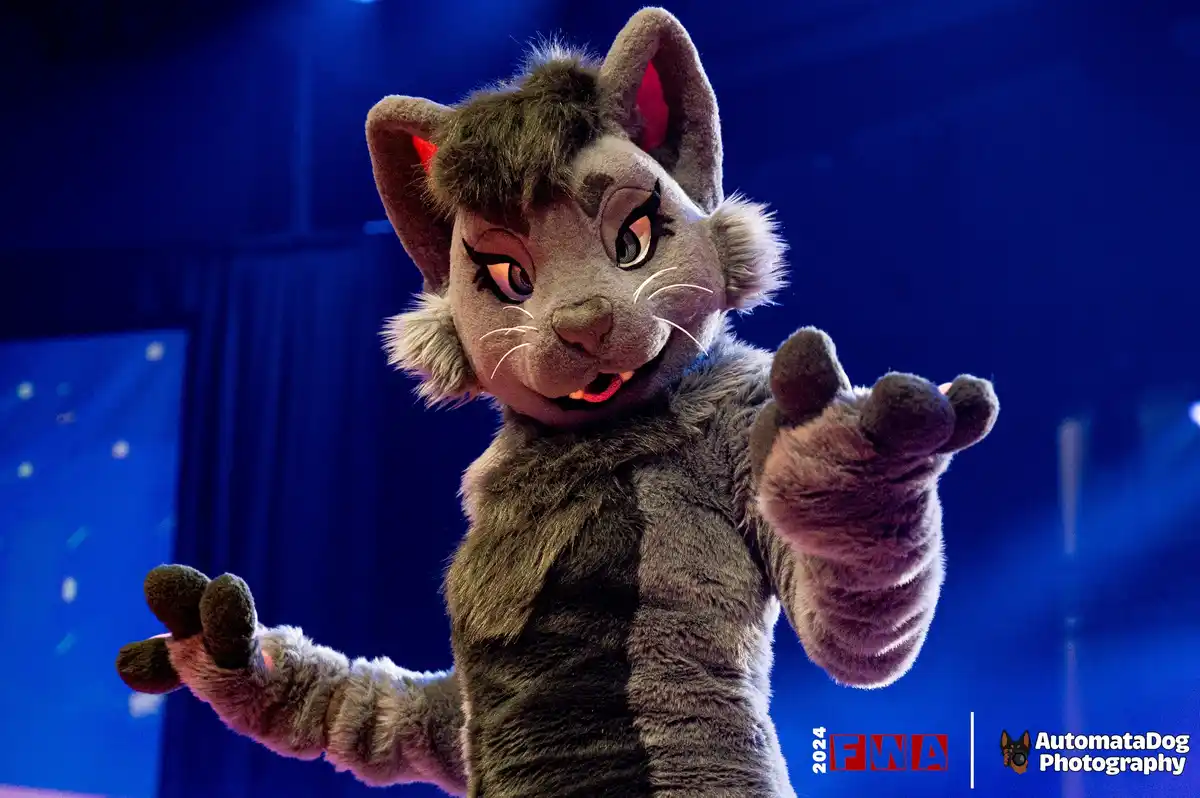 A person in a detailed grey cat fursuit with red inner ears and expressive eyes poses with arms outstretched against a blue…