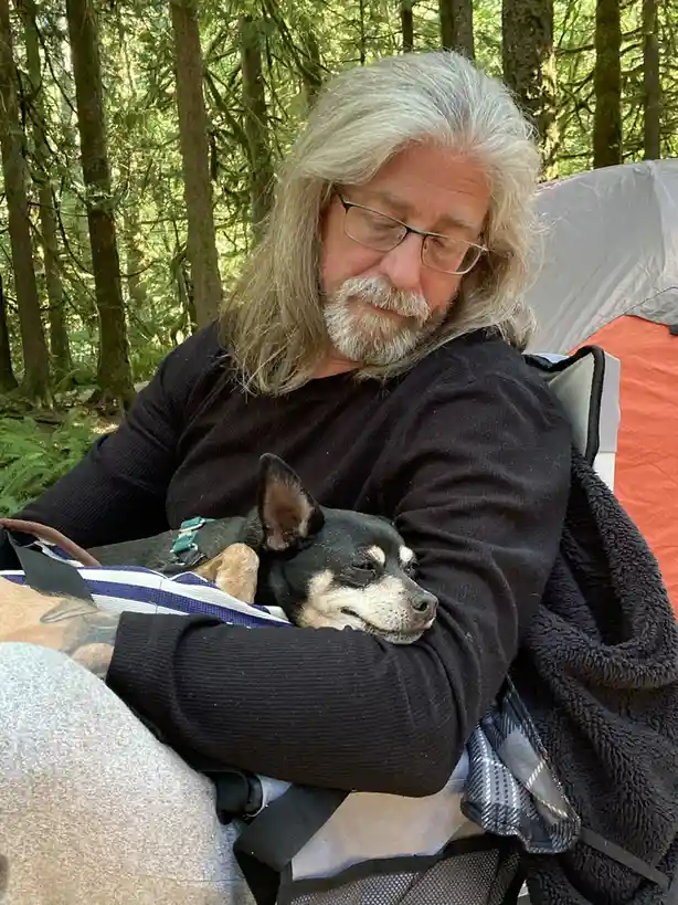 A man with long gray hair and glasses sits in a camping chair in a forest, holding a small black and tan dog that is…