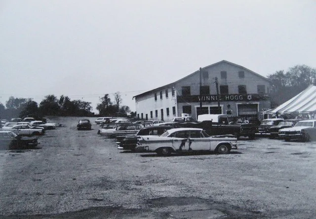 A black-and-white photo shows a vintage car dealership lot filled with classic American cars from the 1950s or 60s. A large…